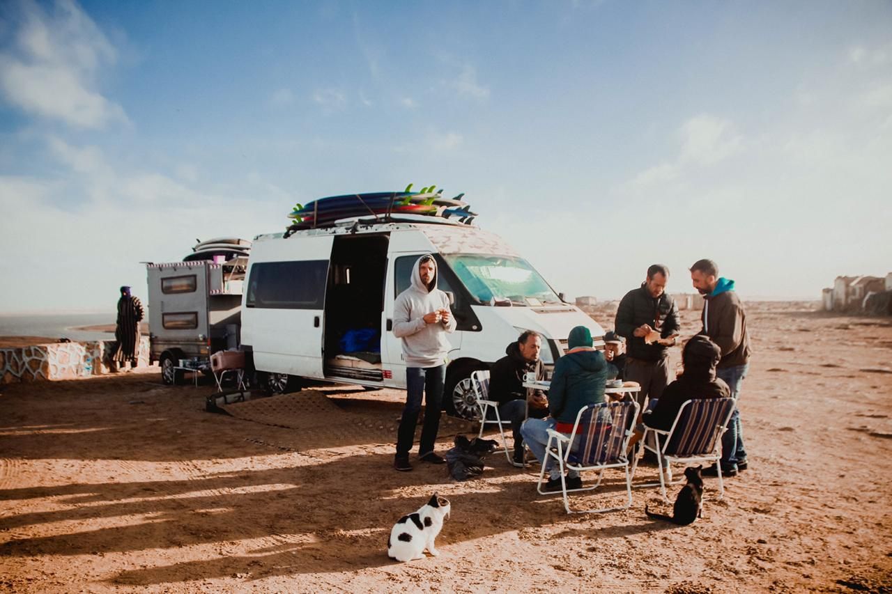Group of people camping near a white van with surfboards on a sandy beach under a blue sky.