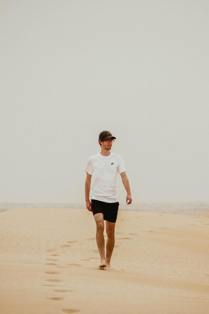 A young man in white Nike t-shirt and black shorts walks barefoot on sandy desert dunes leaving footprints behind.