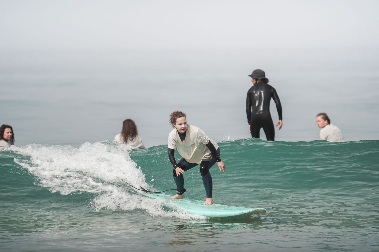 Group of surfers riding waves on a cloudy day, with one smiling surfer on a turquoise board in focus.