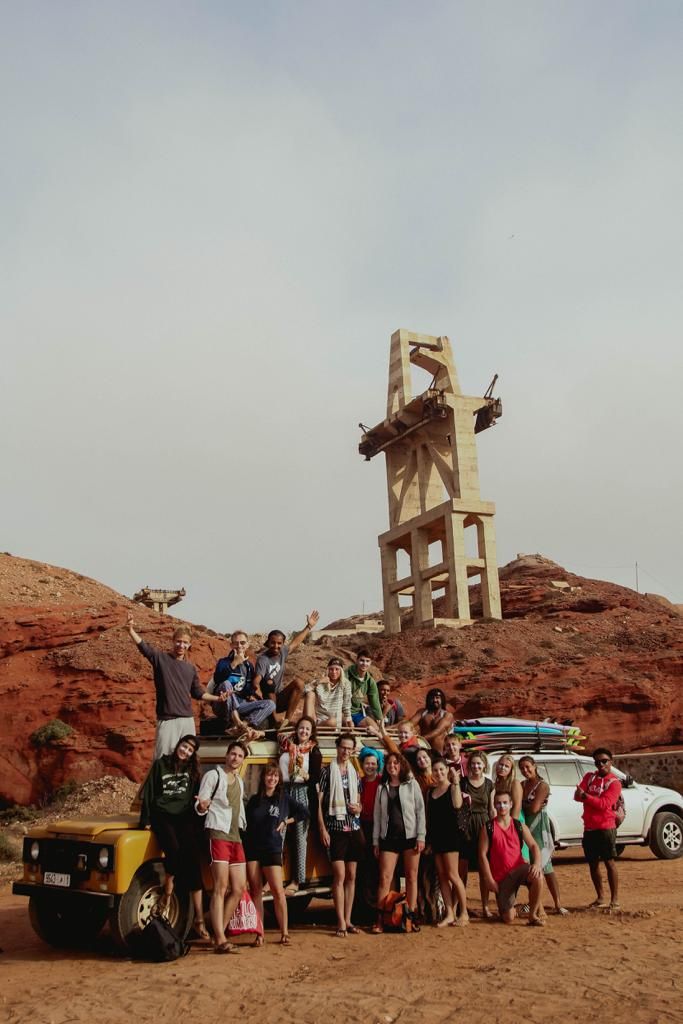 Group of tourists posing near 4x4 vehicles and an abandoned industrial structure in a red desert landscape.