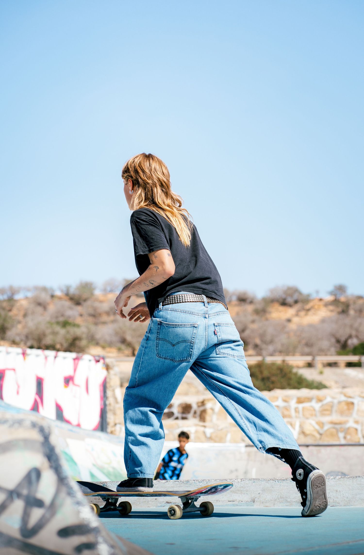 Blonde skateboarder in black shirt and Levis jeans riding skateboard at outdoor skate park.
