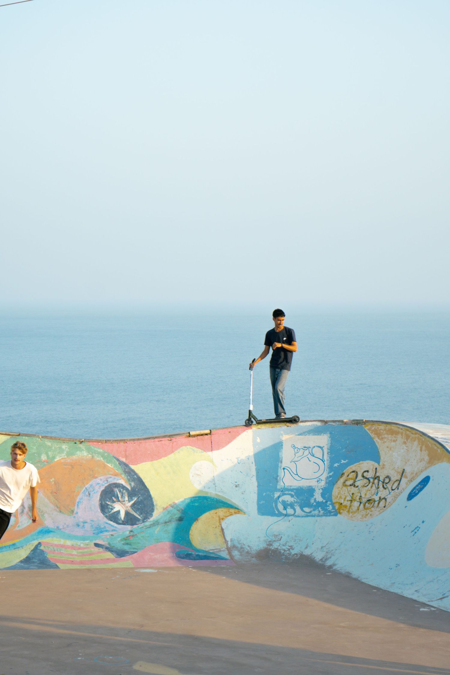 Two youth riders on colorful painted half-pipe ramp with calm ocean visible in misty background.