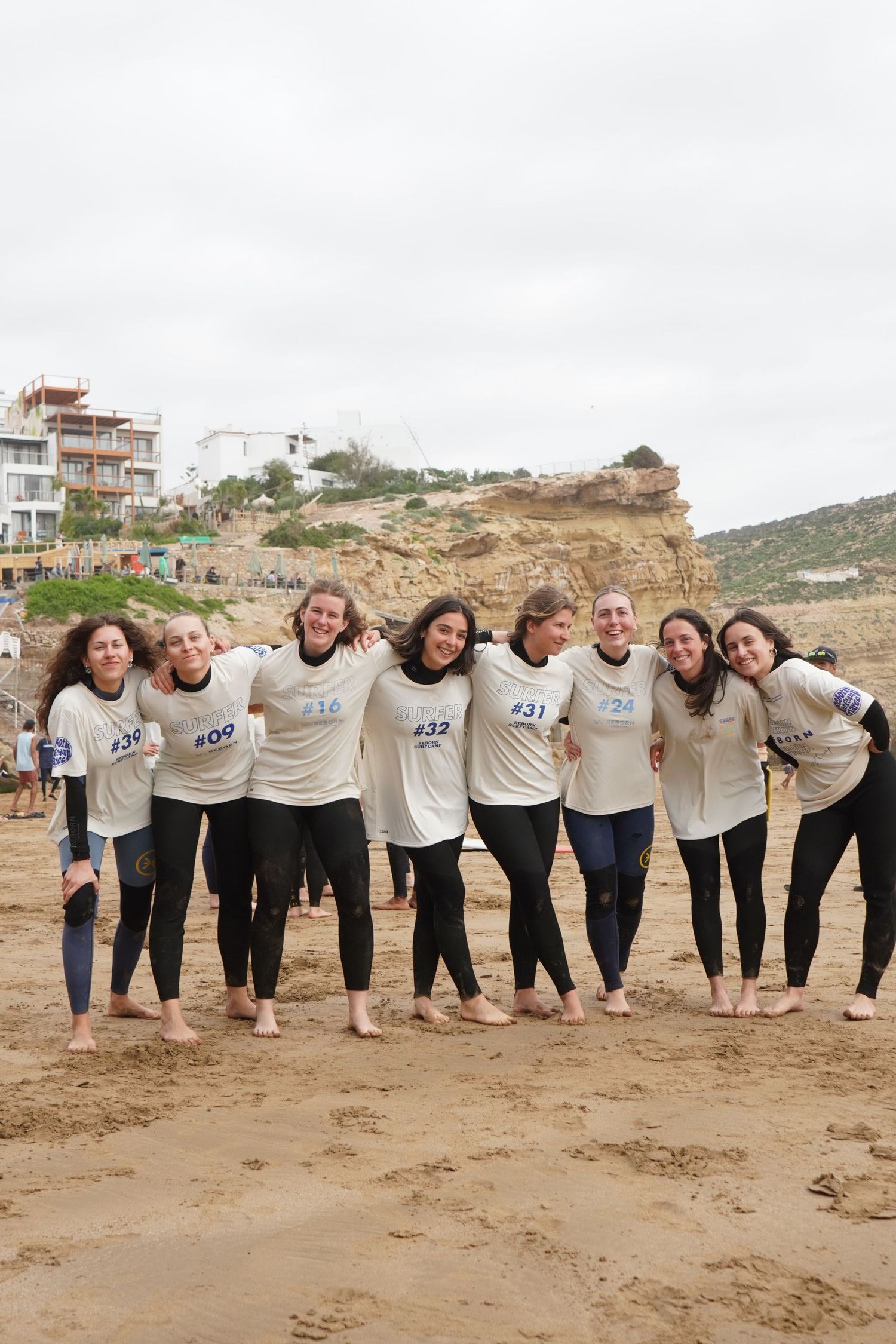 Group of eight women in matching white t-shirts and wetsuits posing together on a sandy beach during a surf competition.