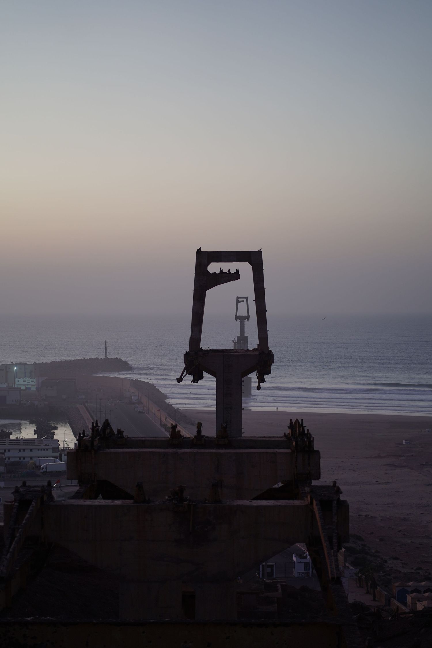 Silhouette of industrial crane structure against a twilight sky overlooking a coastal city beach.