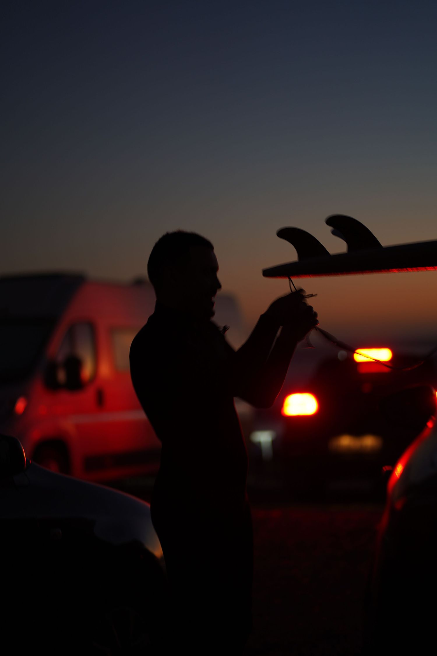 Silhouette of surfer holding board at dusk with red vehicle lights glowing in background.