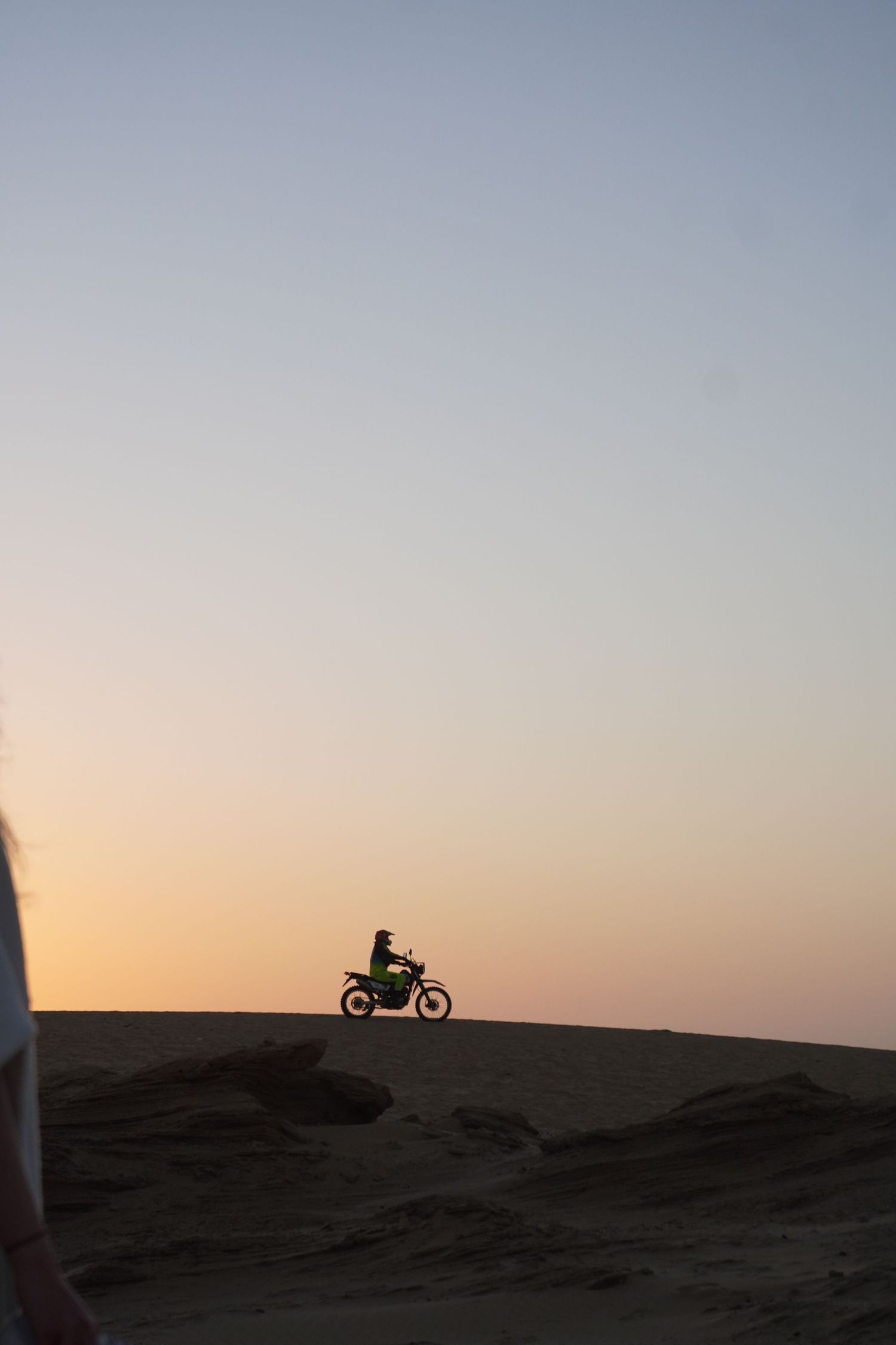 Silhouette of a motorcyclist riding on sand dunes against a warm sunset sky in a desert landscape.