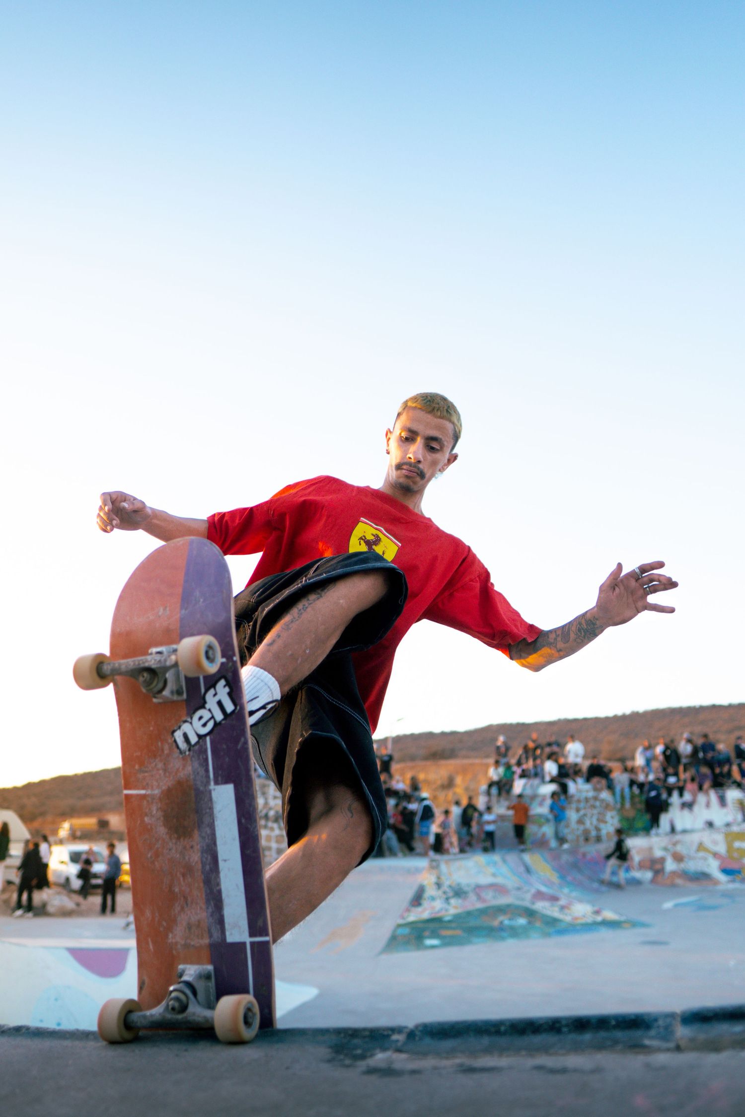 Skateboarder in red Ferrari shirt performs trick at outdoor skate park with crowd watching in background.