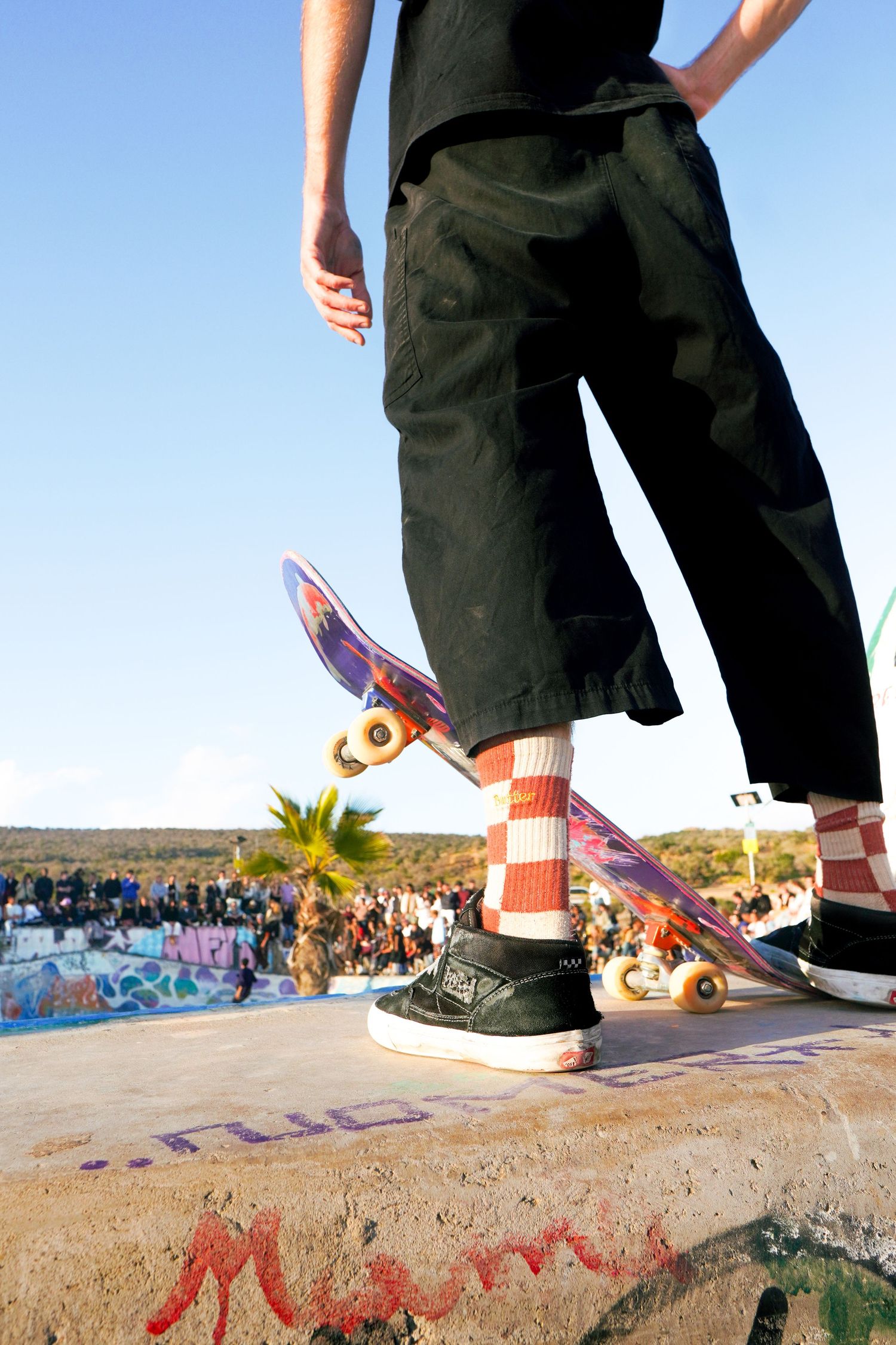 Skateboarder standing on concrete ledge with colorful checkered socks and black skate shoes at outdoor skatepark.