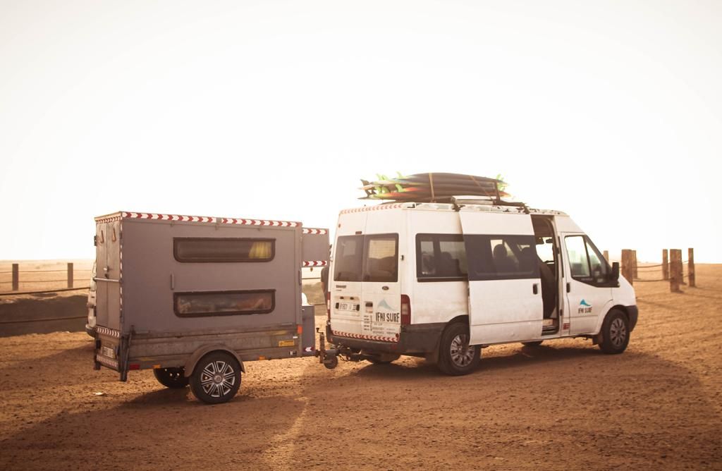 White camper van with rooftop rack towing a gray trailer on a sandy desert road at sunset.