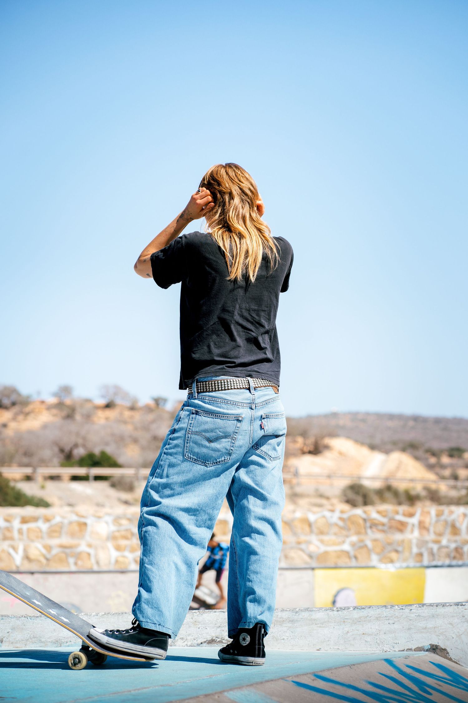 Skateboarder with long blonde hair in baggy jeans standing at outdoor skate park under blue sky.