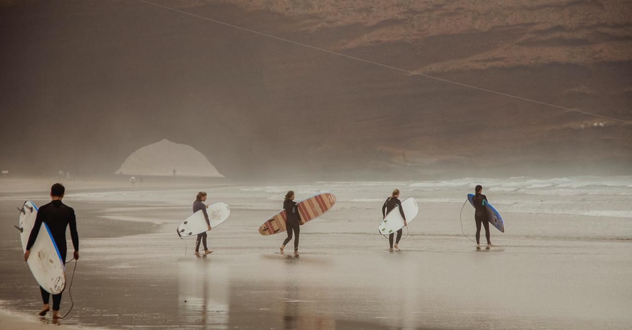 Six surfers carrying boards walk along a misty beach shoreline with dramatic coastal hills in the background.