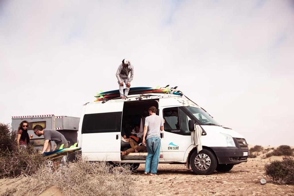 Surfers unload boards from a white van on a sandy beach, with one person sitting on the roof rack.