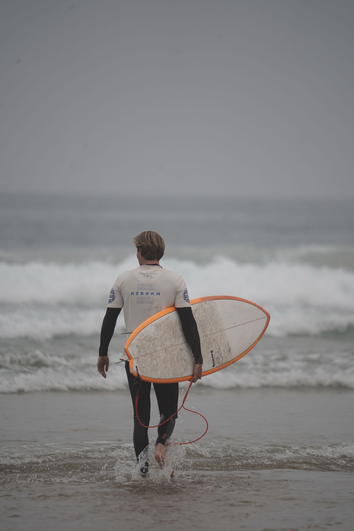 Surfer in wetsuit carrying white surfboard with orange rails walks toward ocean waves on overcast day.