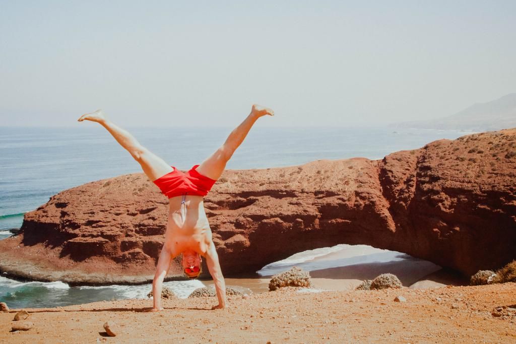 Person doing a handstand on sandy beach near red rock arch formation with ocean backdrop.