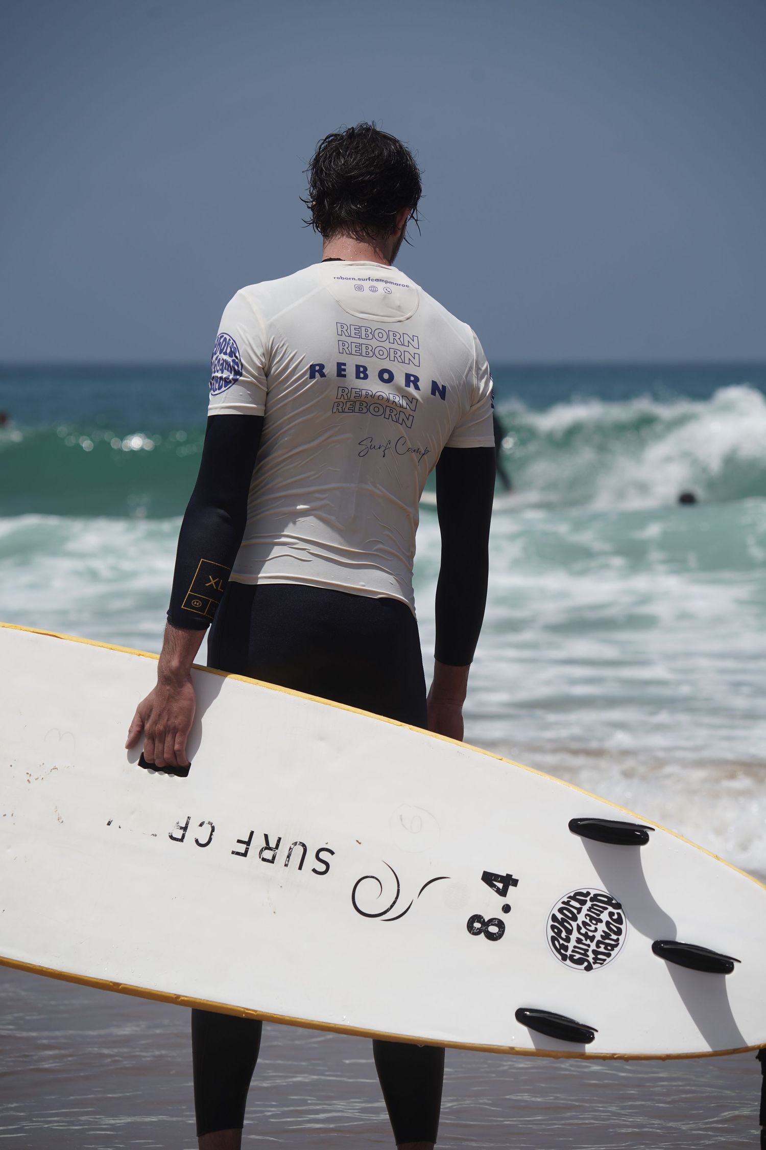 A surfer in a white Reborn rash guard holds an 8.4 surfboard while standing on the beach facing ocean waves.