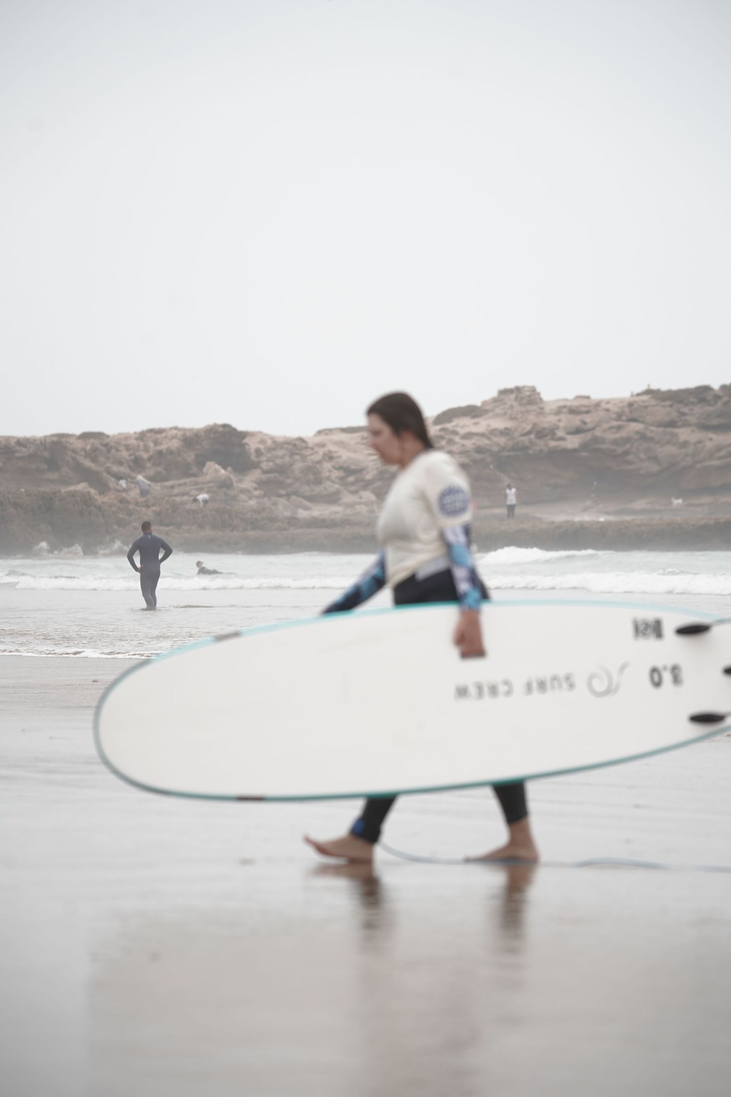 Woman in wetsuit carrying a white surfboard on a misty beach with rocky coastline in the background.