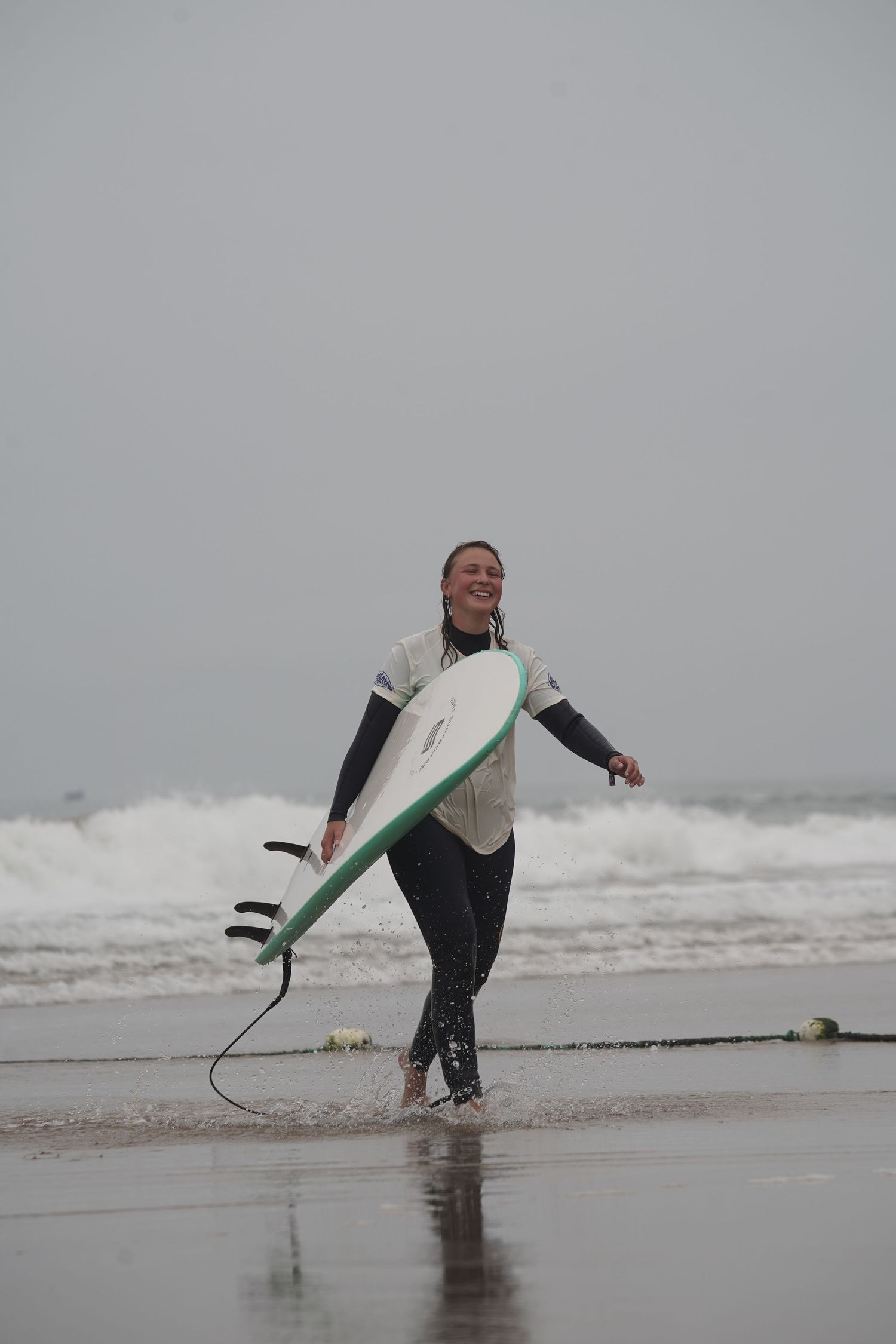 Smiling female surfer in wetsuit carrying white and green surfboard while walking on a misty beach.