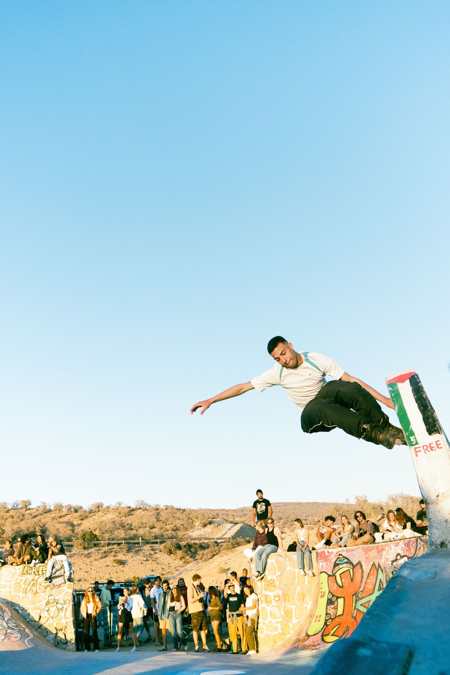 Skateboarder launching high aerial jump over graffiti wall with Palestinian flag visible at outdoor skatepark.