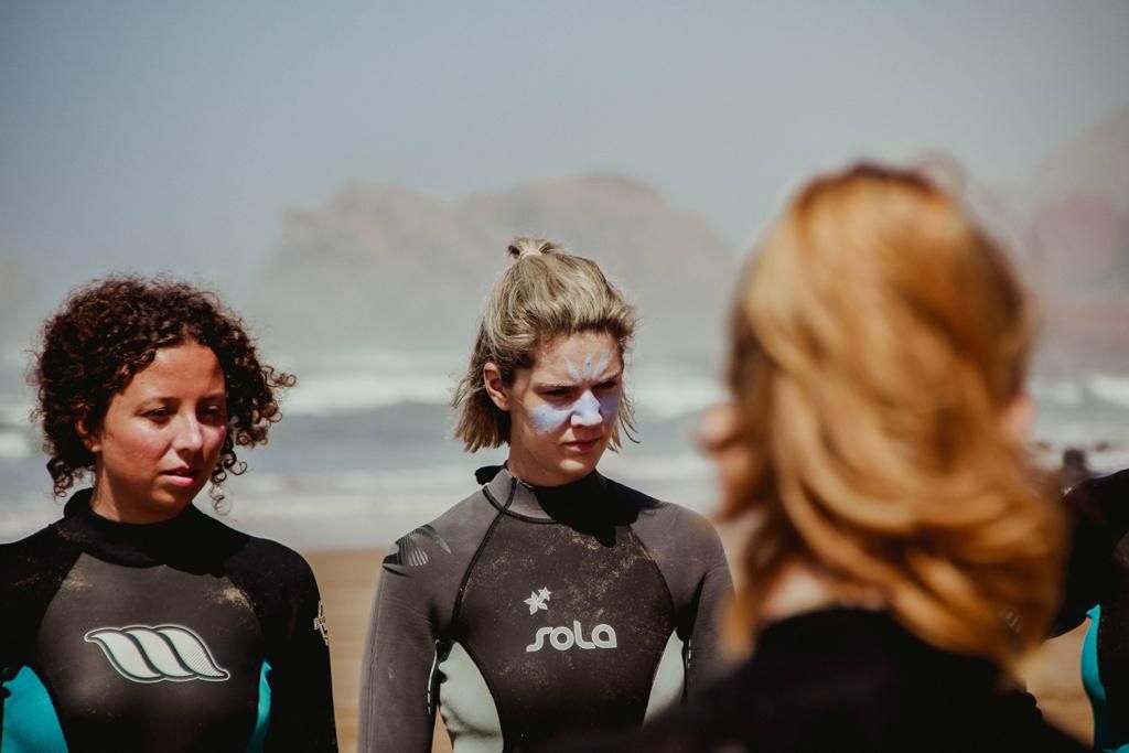 Two women in Sola wetsuits stand on a beach with rocky cliffs in the background during a surf lesson.