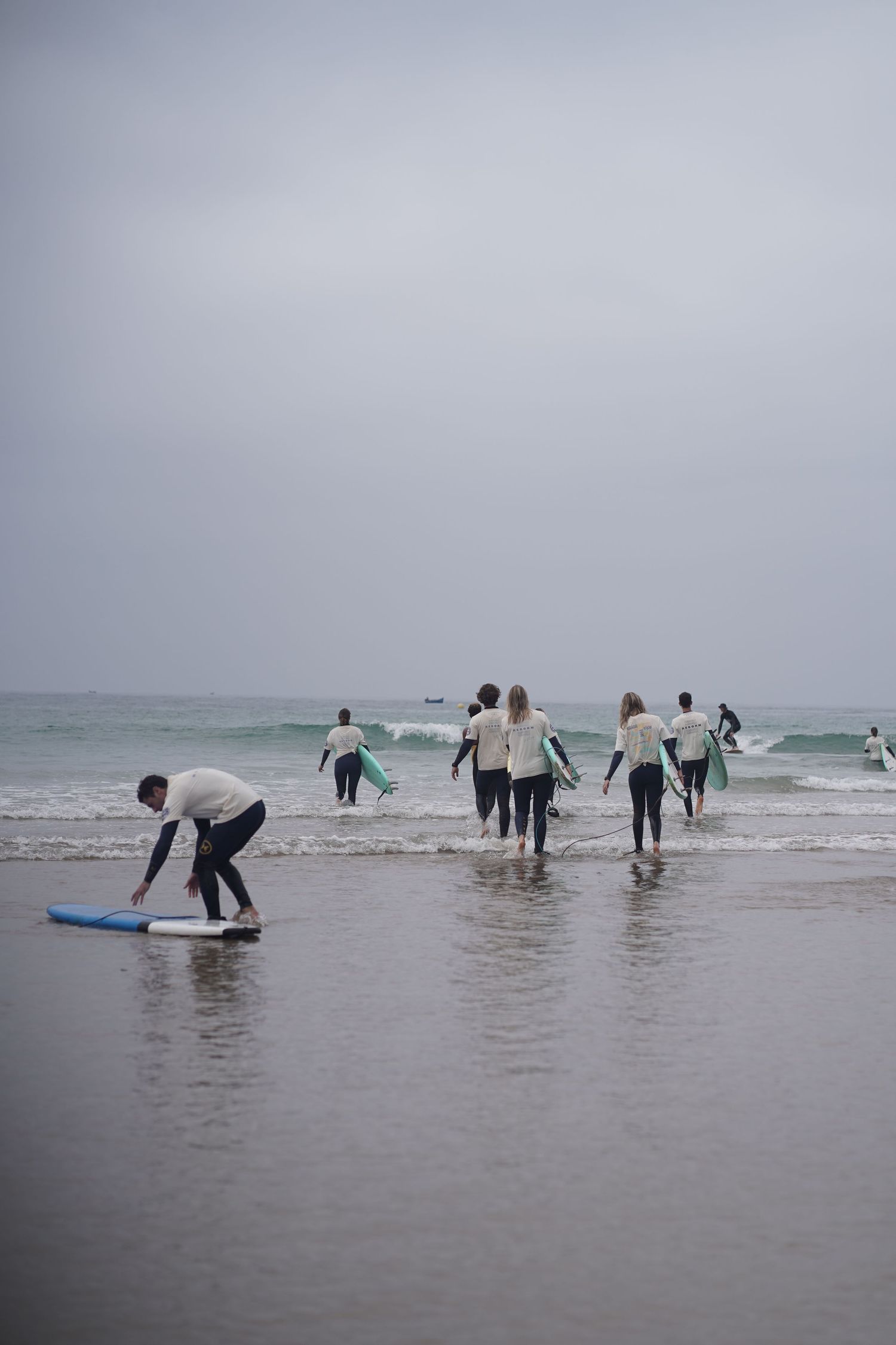Group of surfers in wetsuits carrying boards into ocean waves on a cloudy day at a surf lesson.