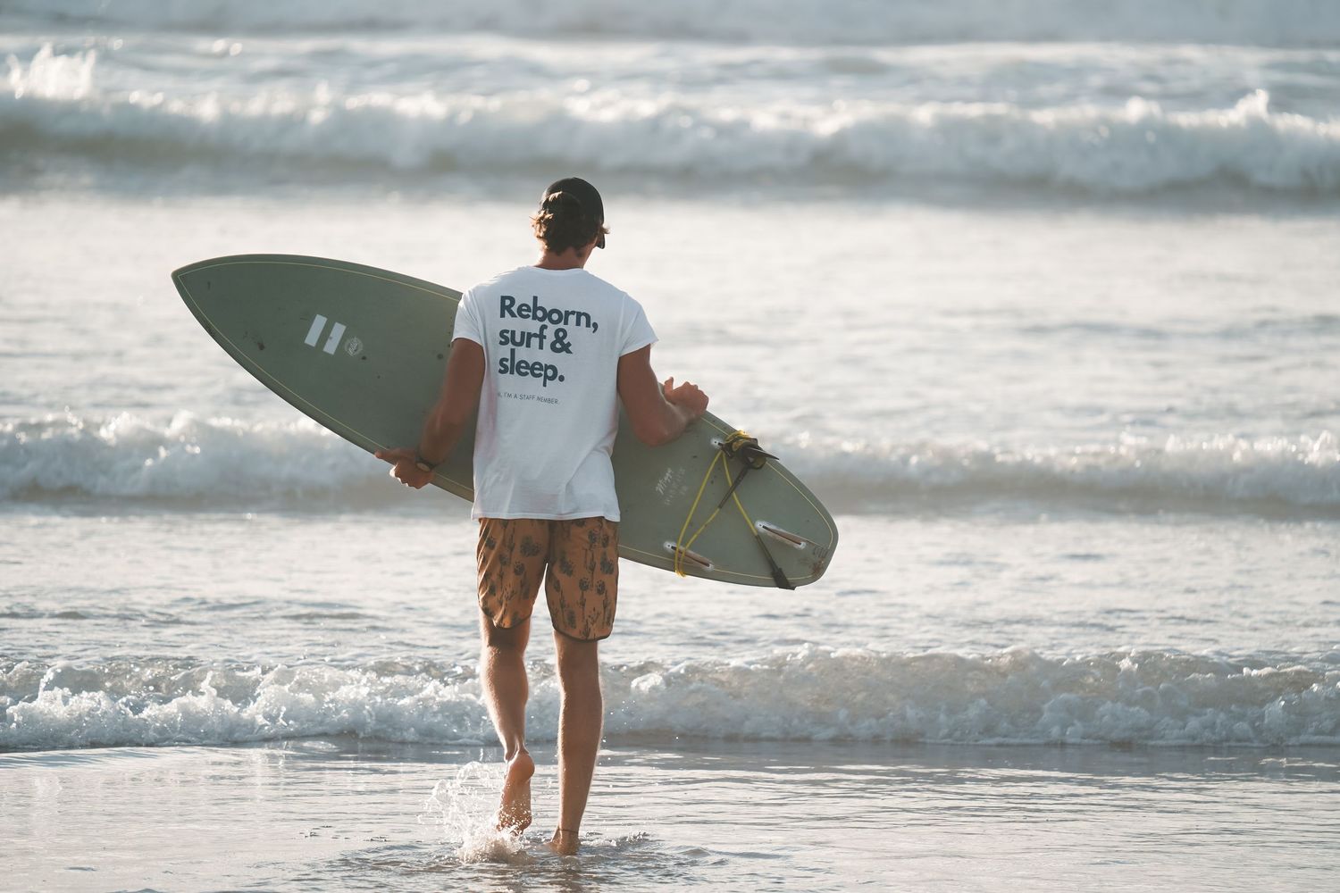 Surfer carrying a green surfboard walking into ocean waves wearing a white t-shirt and floral shorts on a sunny beach.