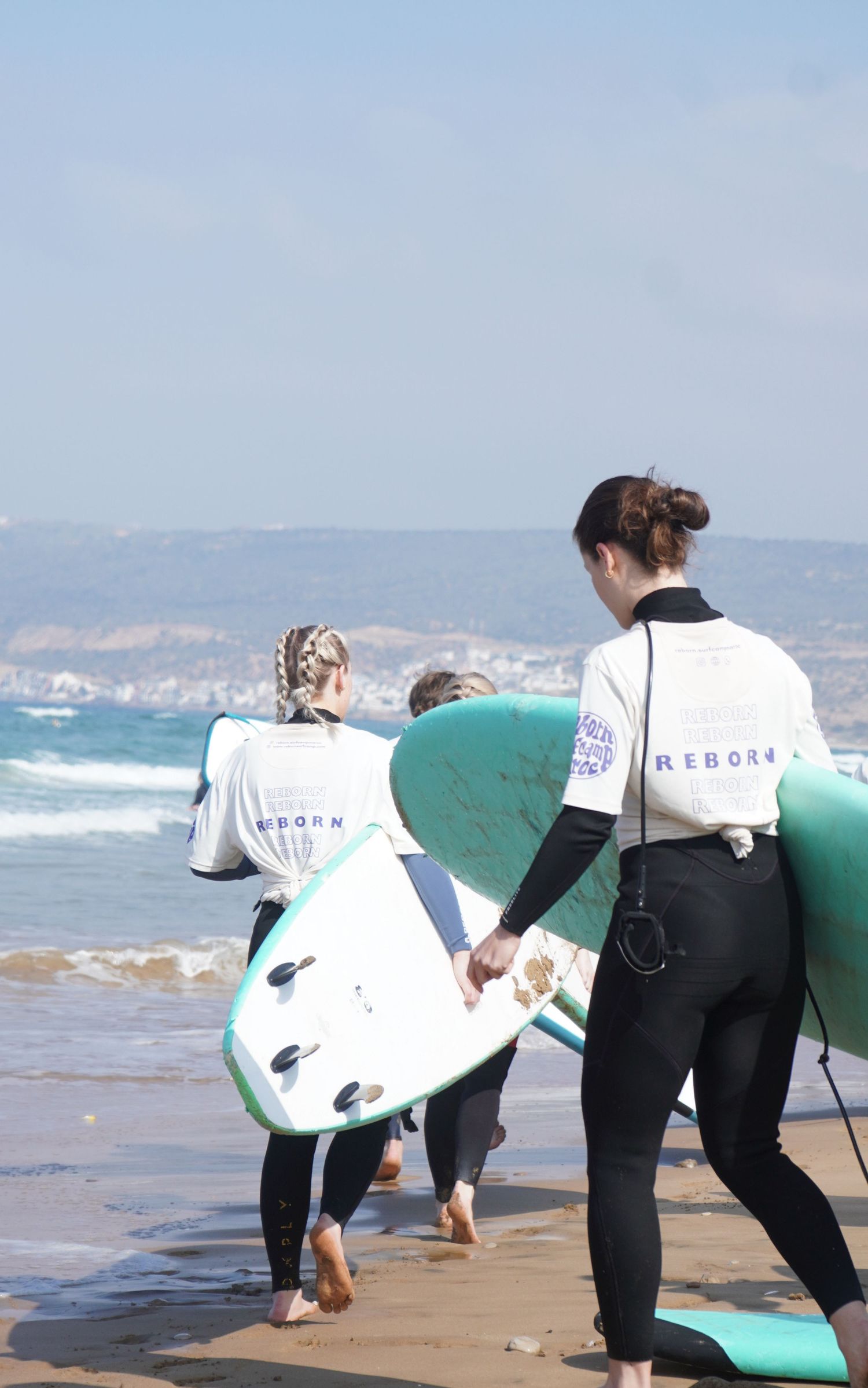 Three surfers in Reborn wetsuits carrying surfboards toward ocean waves on a sandy beach.