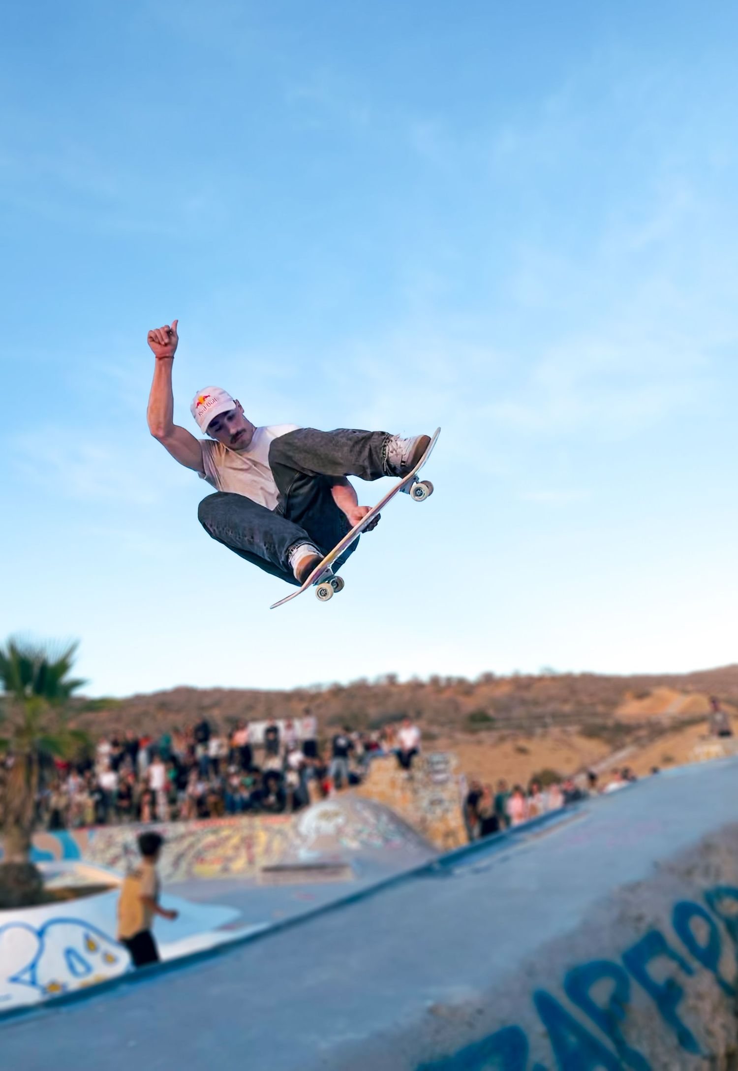 Skateboarder performing a high aerial trick at an outdoor skate park with a crowd watching.