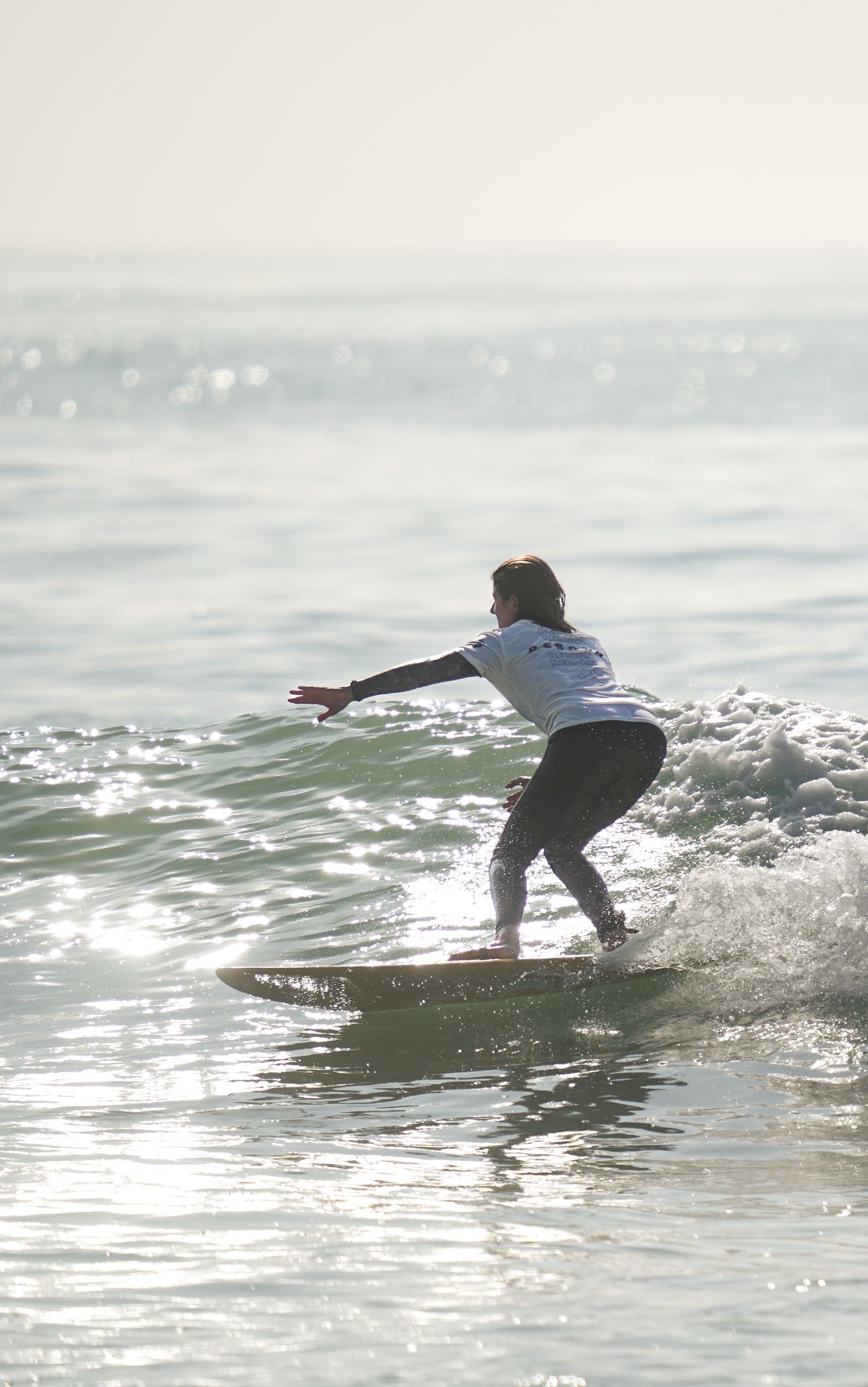 Surfer riding a small wave in the ocean, wearing a white shirt and black pants, with arms outstretched for balance.
