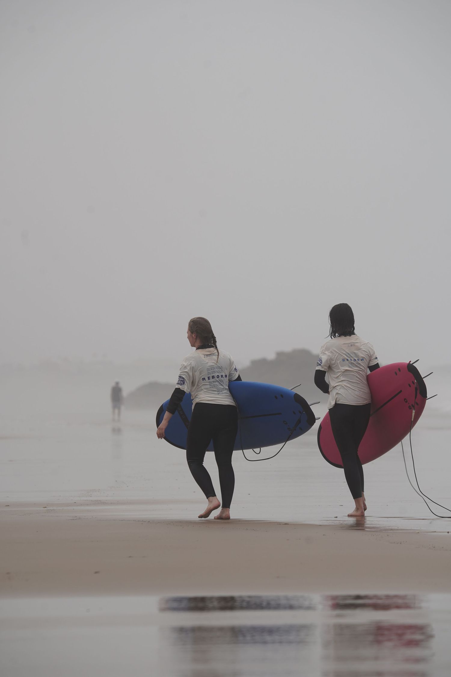 Two surfers in wetsuits carry blue and pink surfboards along a misty beach toward the ocean.