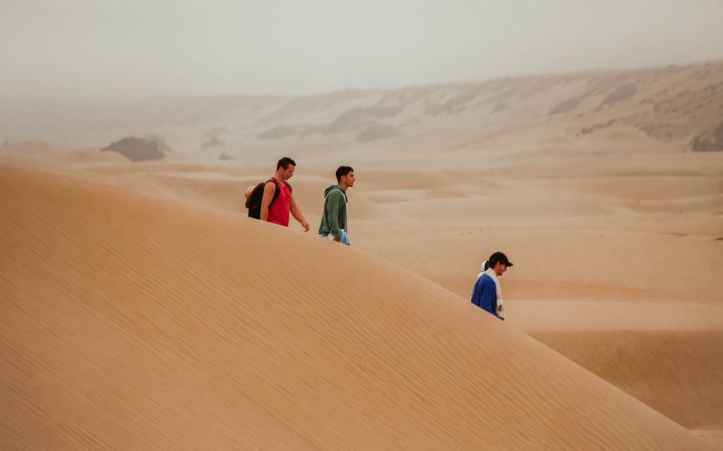 Three people hiking across golden sand dunes in a vast desert landscape under a hazy sky.