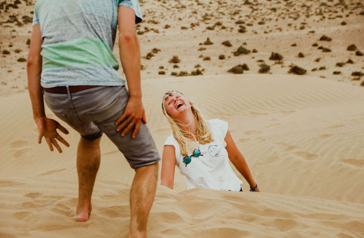A laughing blonde woman sits on a sandy desert dune while a man in gray shorts stands nearby.