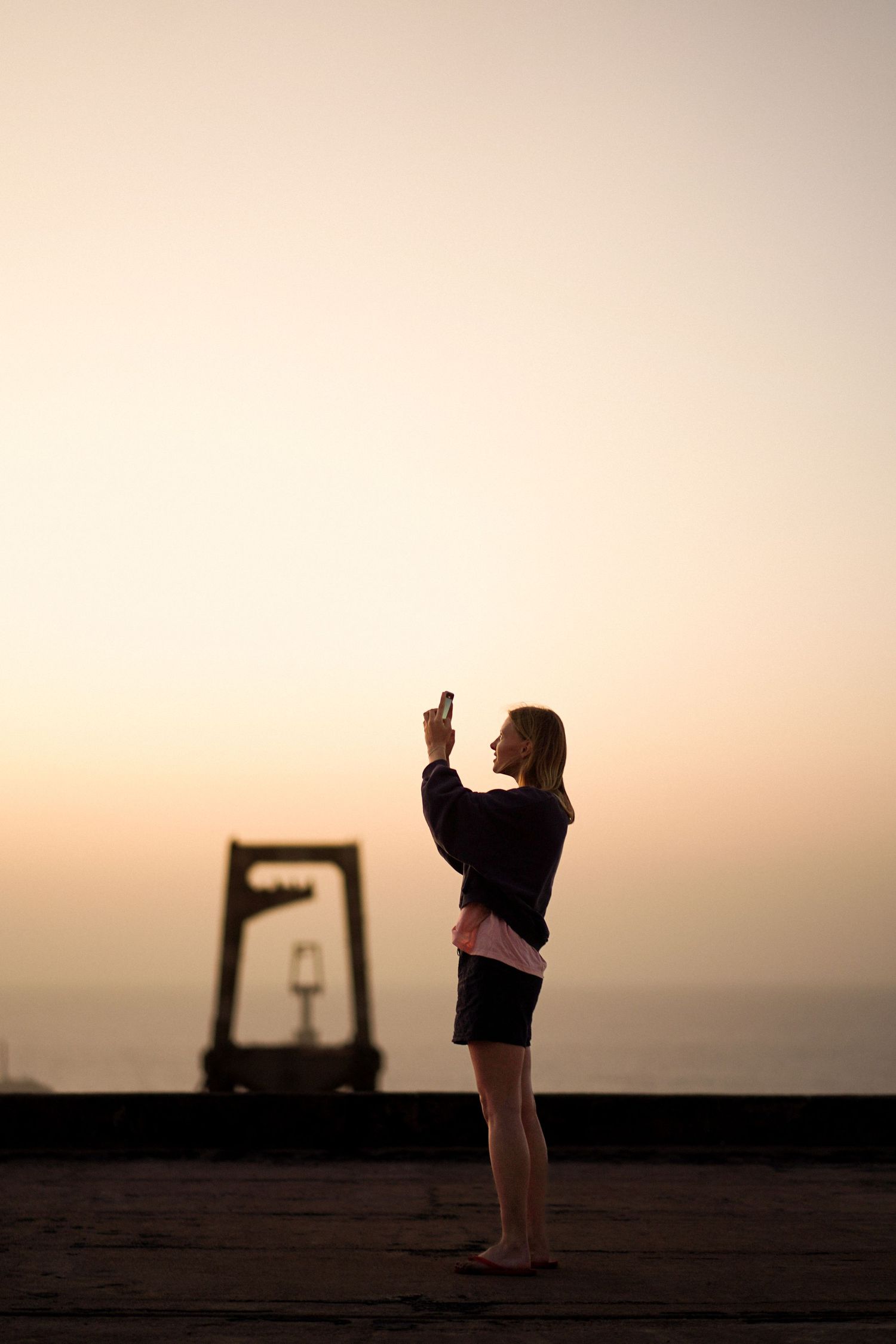 Woman silhouetted against a warm sunset sky, standing on a rooftop taking a photo with her smartphone.