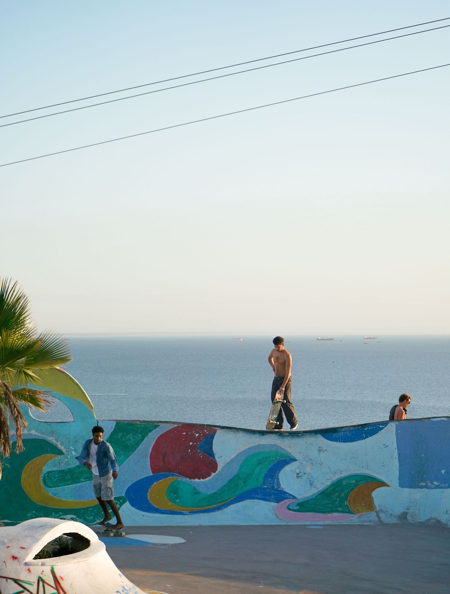 Skateboarders at a coastal skate park with colorful murals and ocean horizon visible in the background.