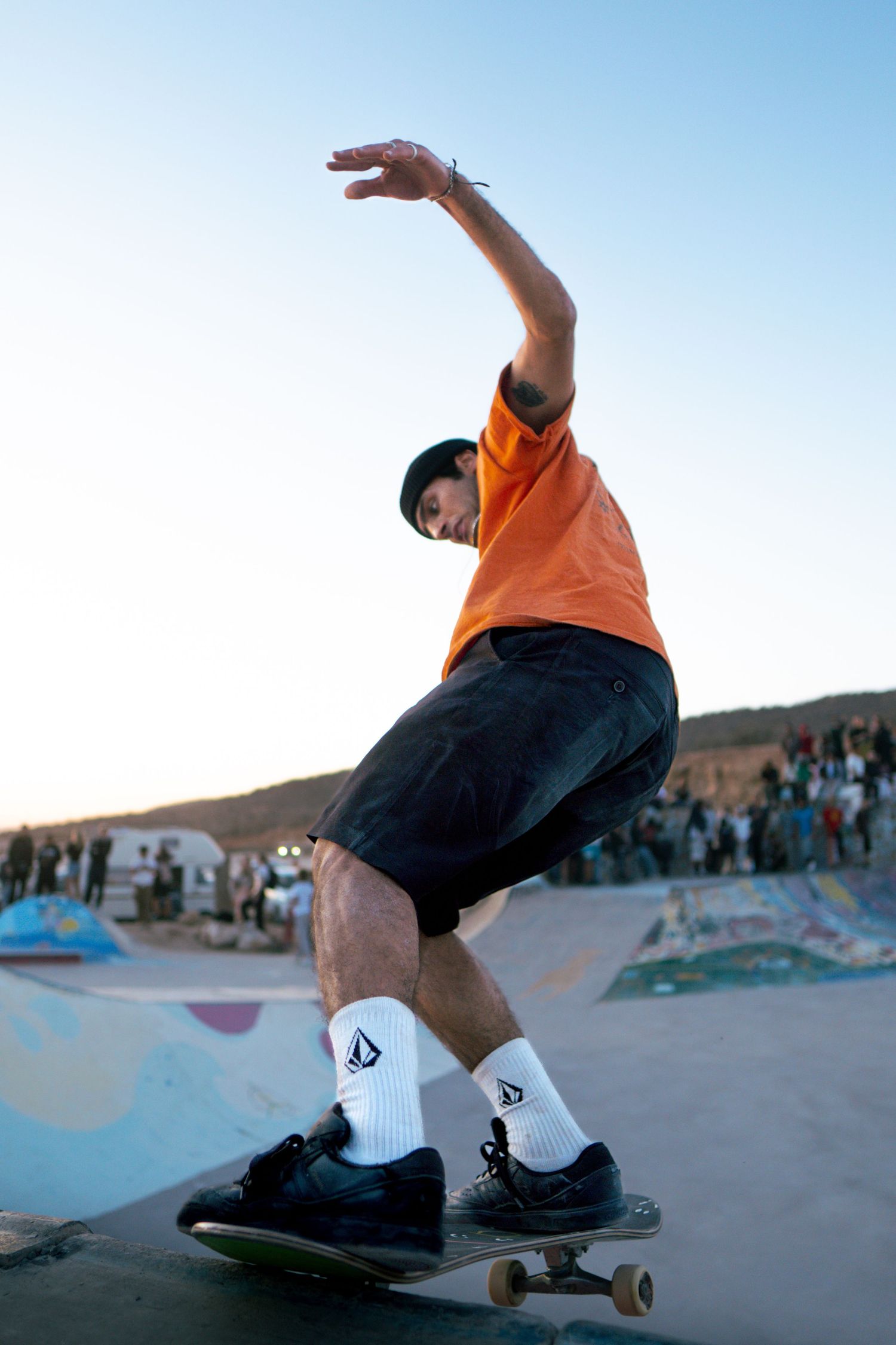 Skateboarder in orange shirt and black cap balances on skateboard at colorful outdoor skate park at sunset.
