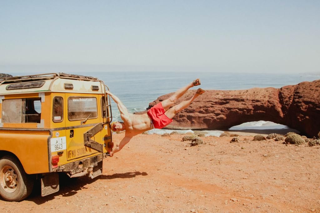 Person doing a dramatic dive pose beside a vintage yellow Land Rover on red desert terrain near ocean arch rock.