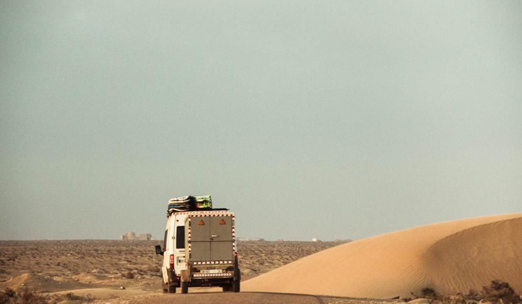 A white camper van drives along a desert road surrounded by vast sandy dunes under a hazy sky.