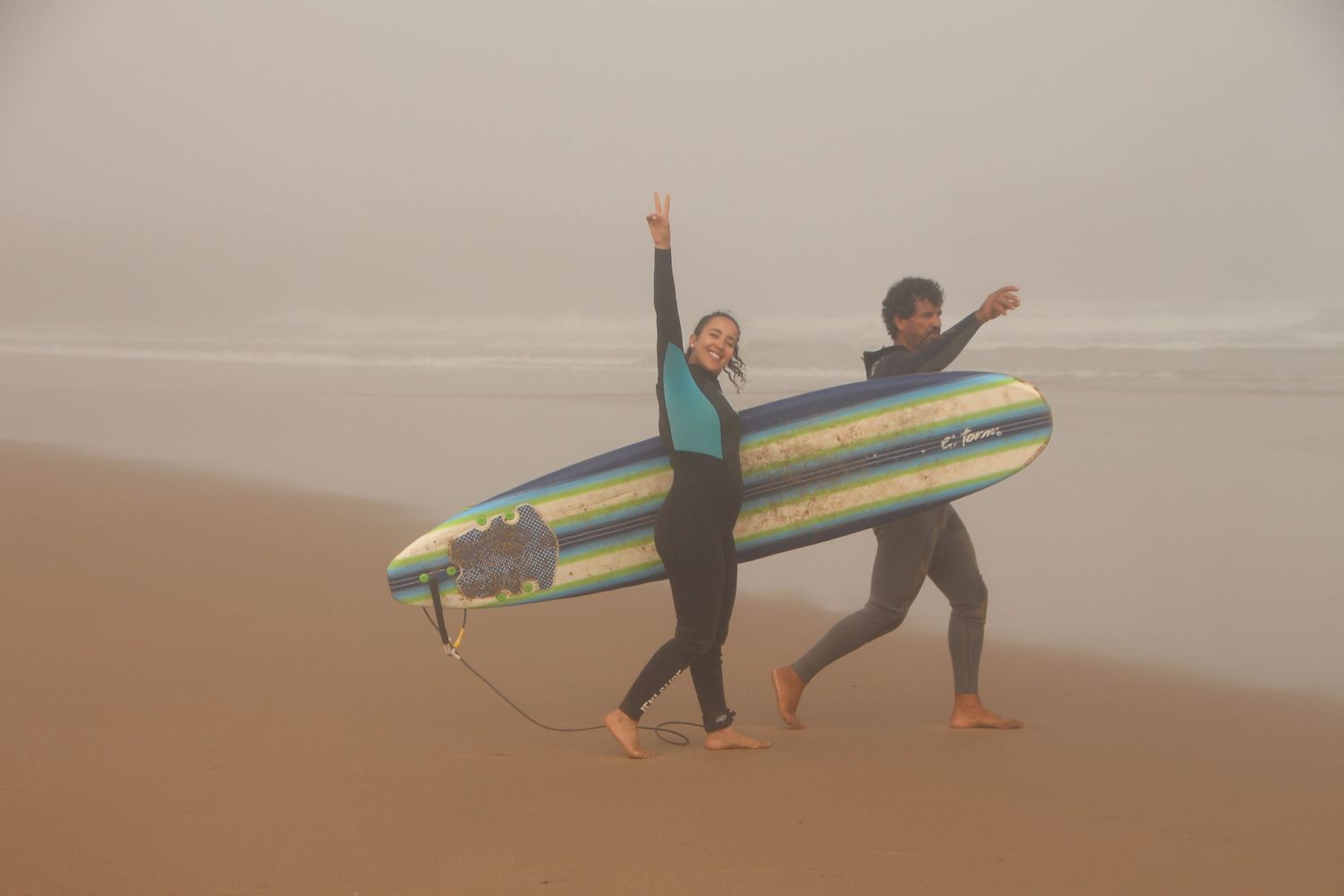 Two surfers in wetsuits carry a colorful surfboard on a misty beach, celebrating with raised arms.