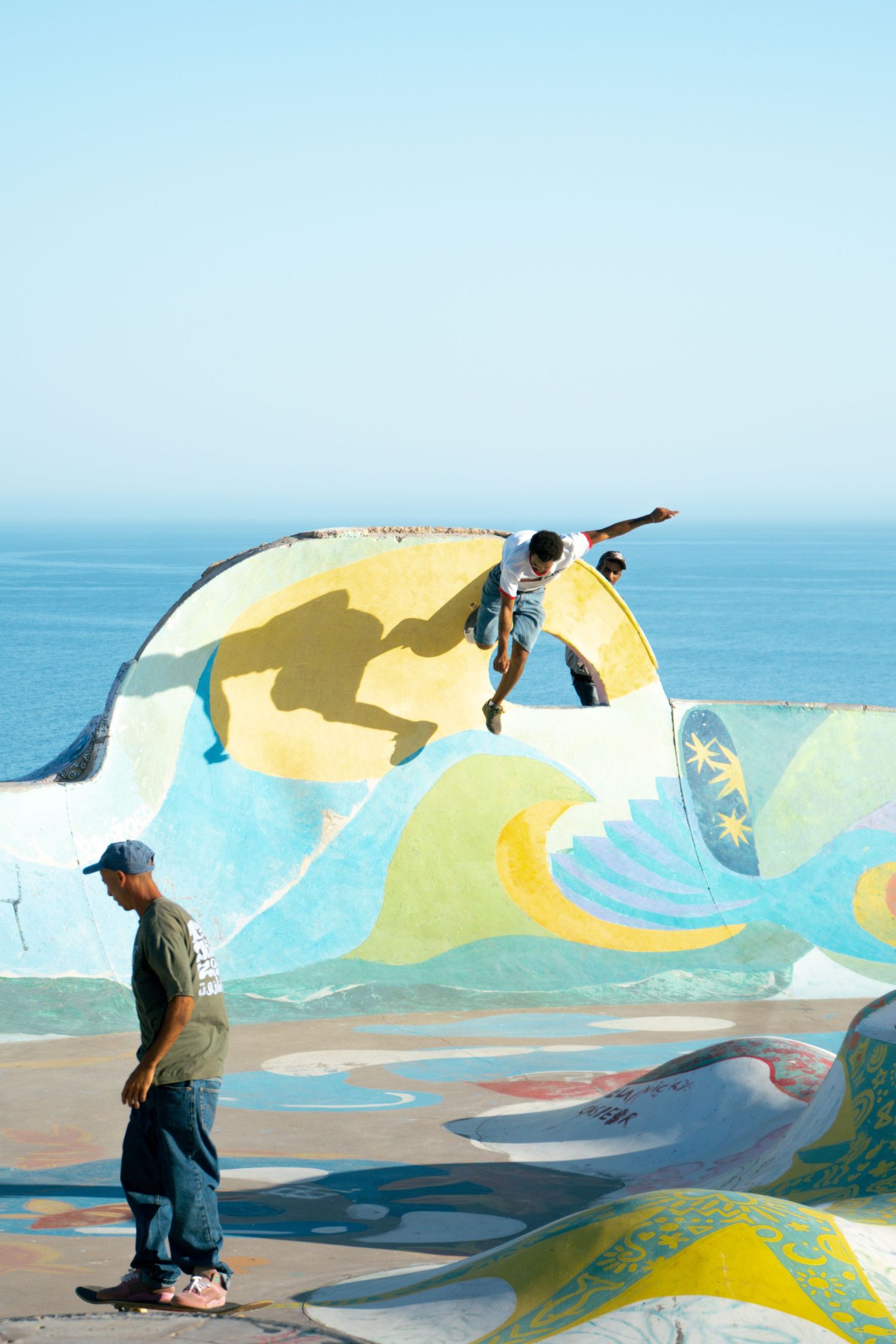 Skateboarder soars above colorful wave-painted ramp at oceanside skate park while another skater watches below.