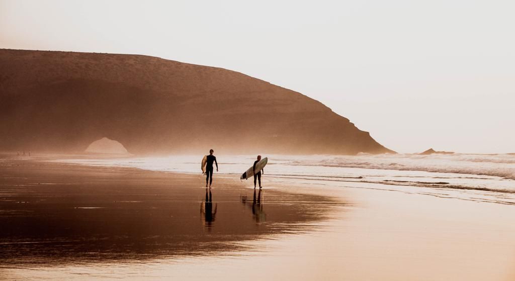 Two surfers walk along a misty beach at golden hour with surfboards, rocky cliffs in the background.