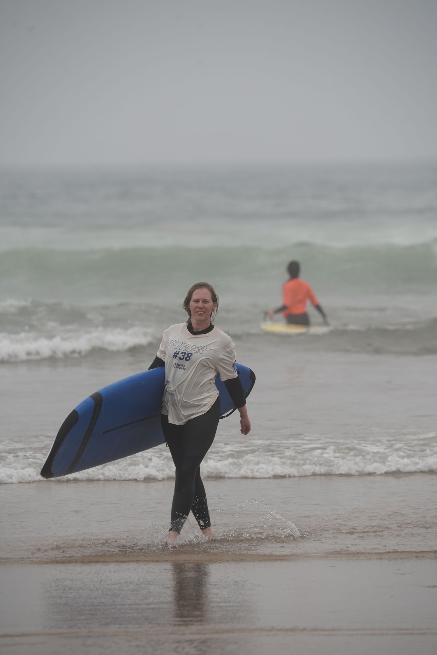 Woman in wetsuit carrying a blue surfboard walking out of ocean waves on a misty beach day.