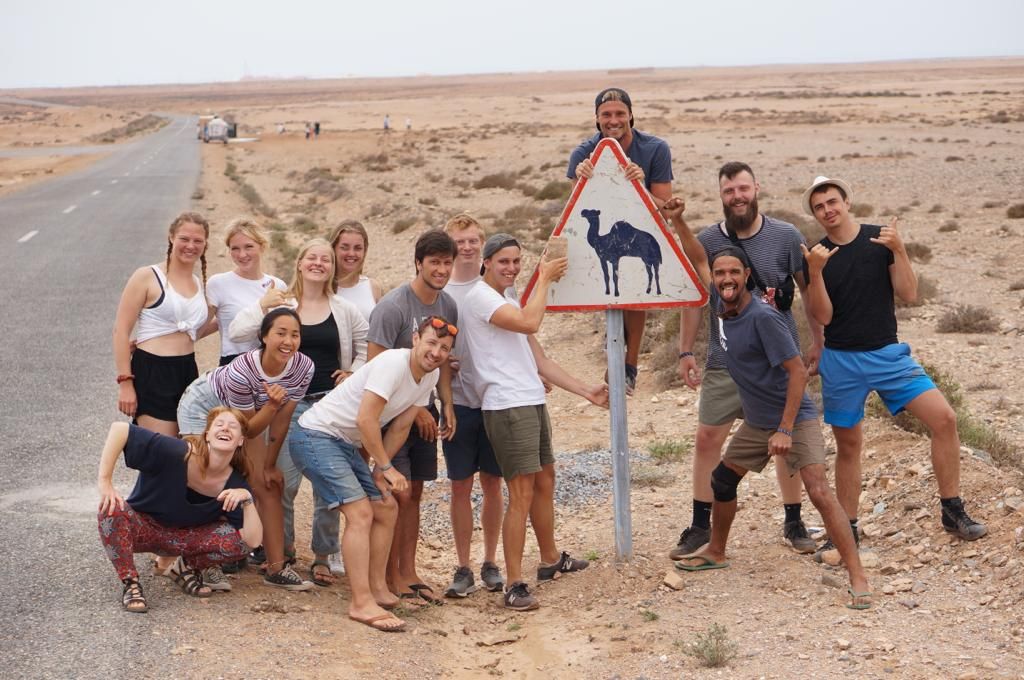Group of tourists posing playfully around a camel warning road sign in a desert landscape.