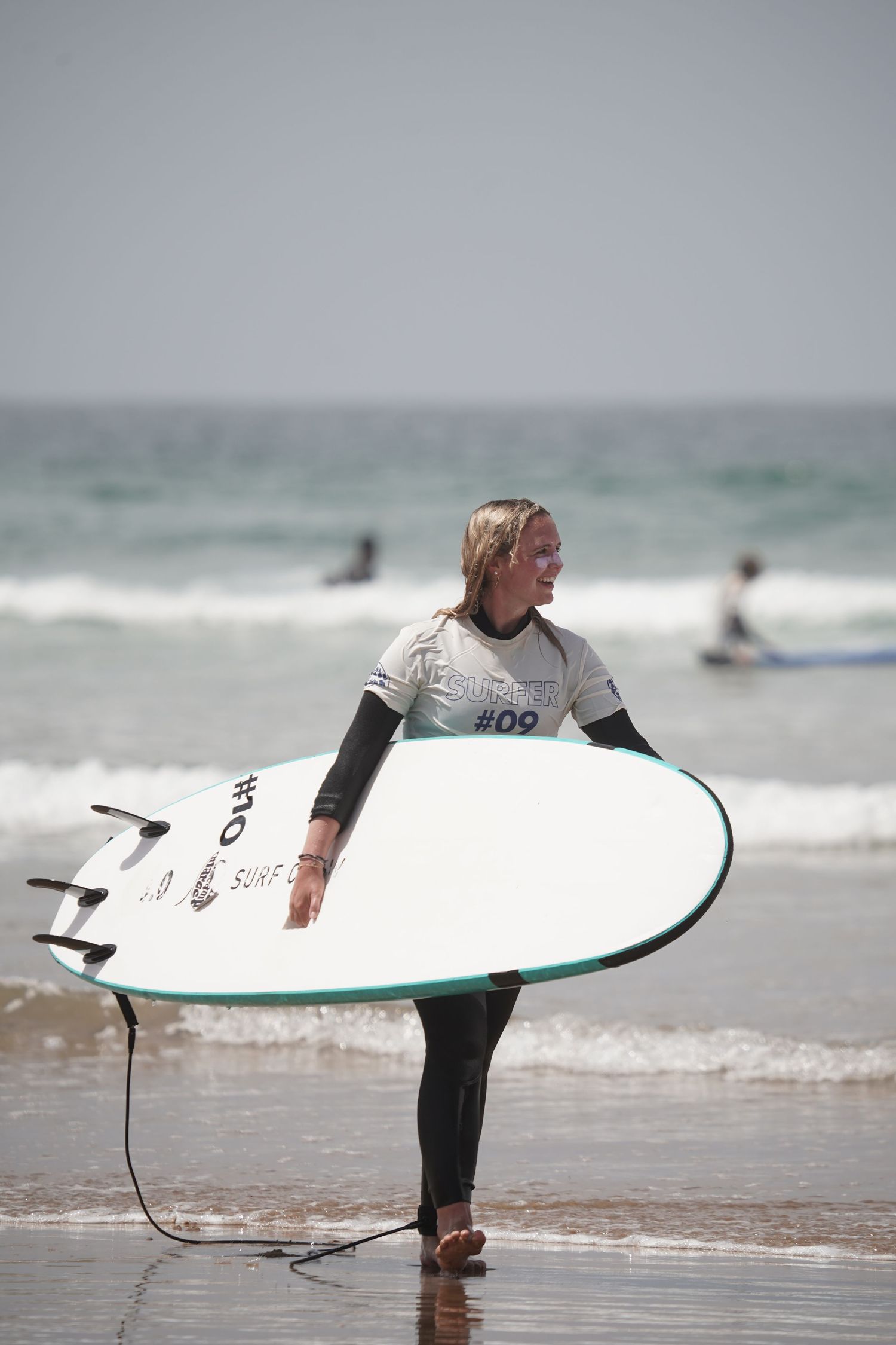 Female surfer in wetsuit carrying white surfboard numbered 09 walks along sandy beach with ocean waves behind her.