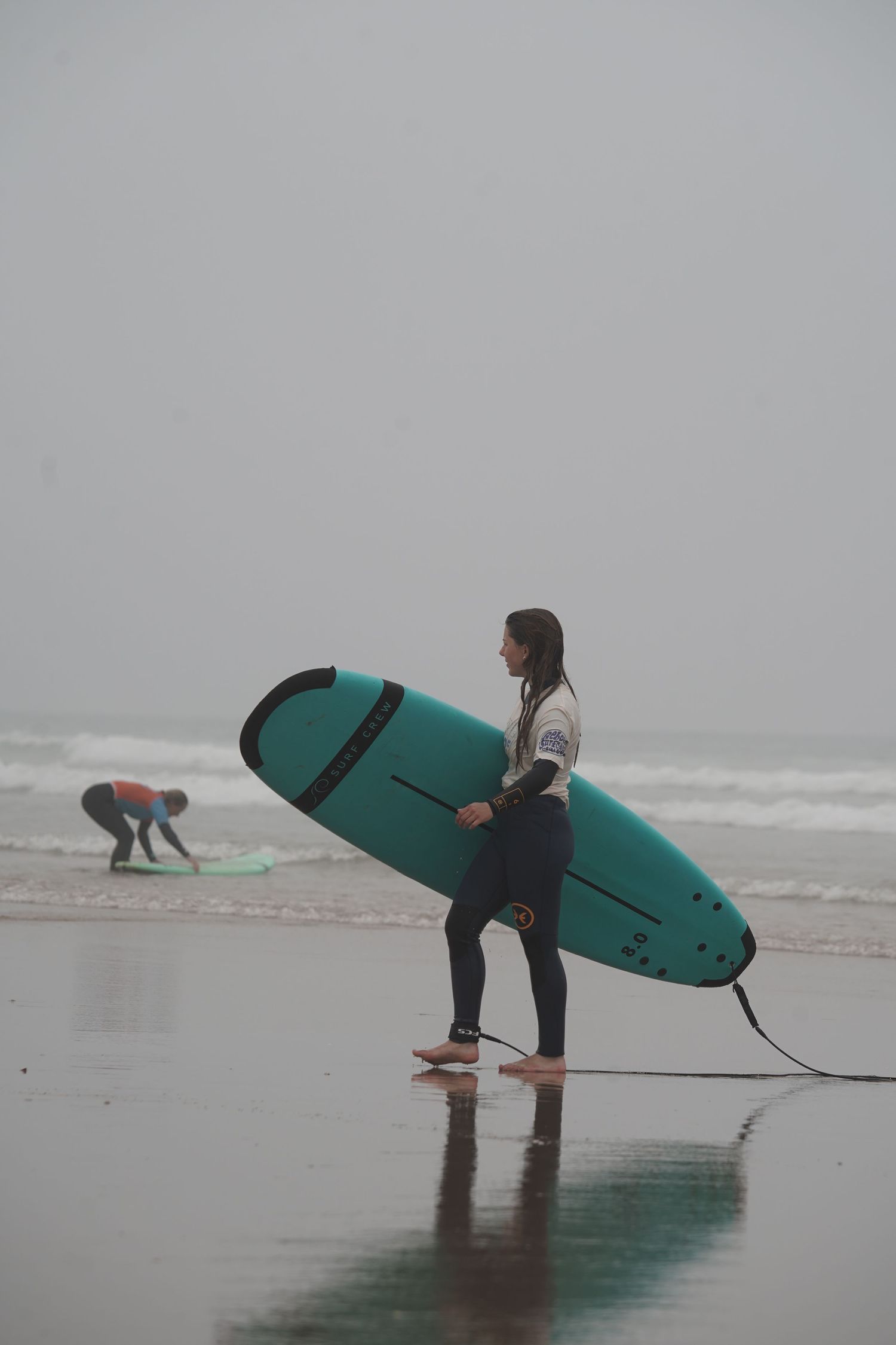 A woman in a wetsuit carries a teal surfboard on a misty beach while another surfer prepares in the background.