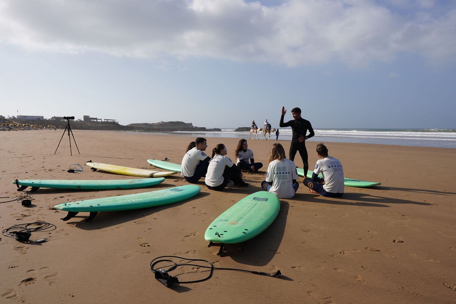 Surf instructor teaching a group of students on a sandy beach with colorful surfboards laid out in front of them.