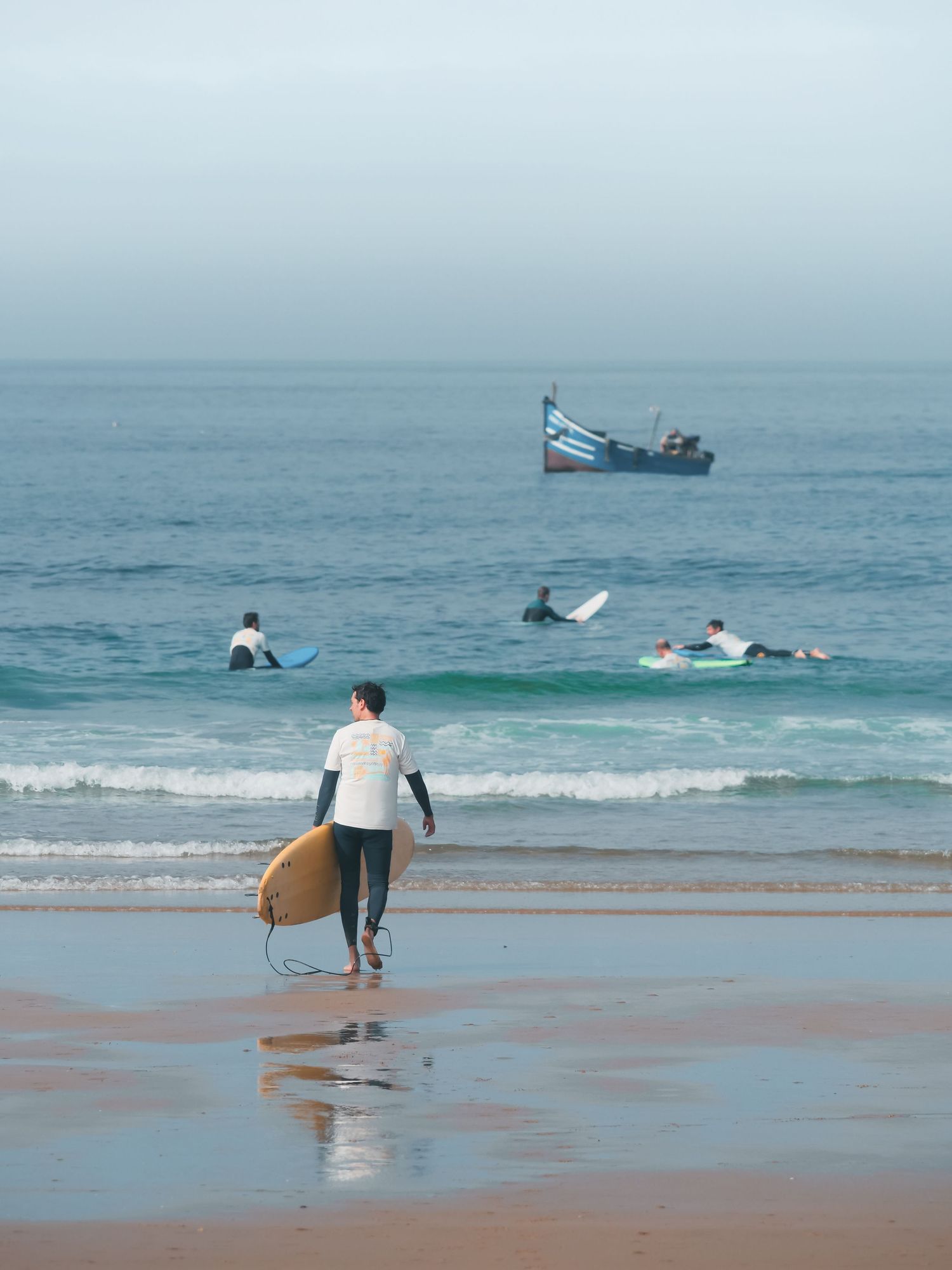 Surfer carrying a yellow board walks toward ocean waves with other surfers and a boat visible in the background.