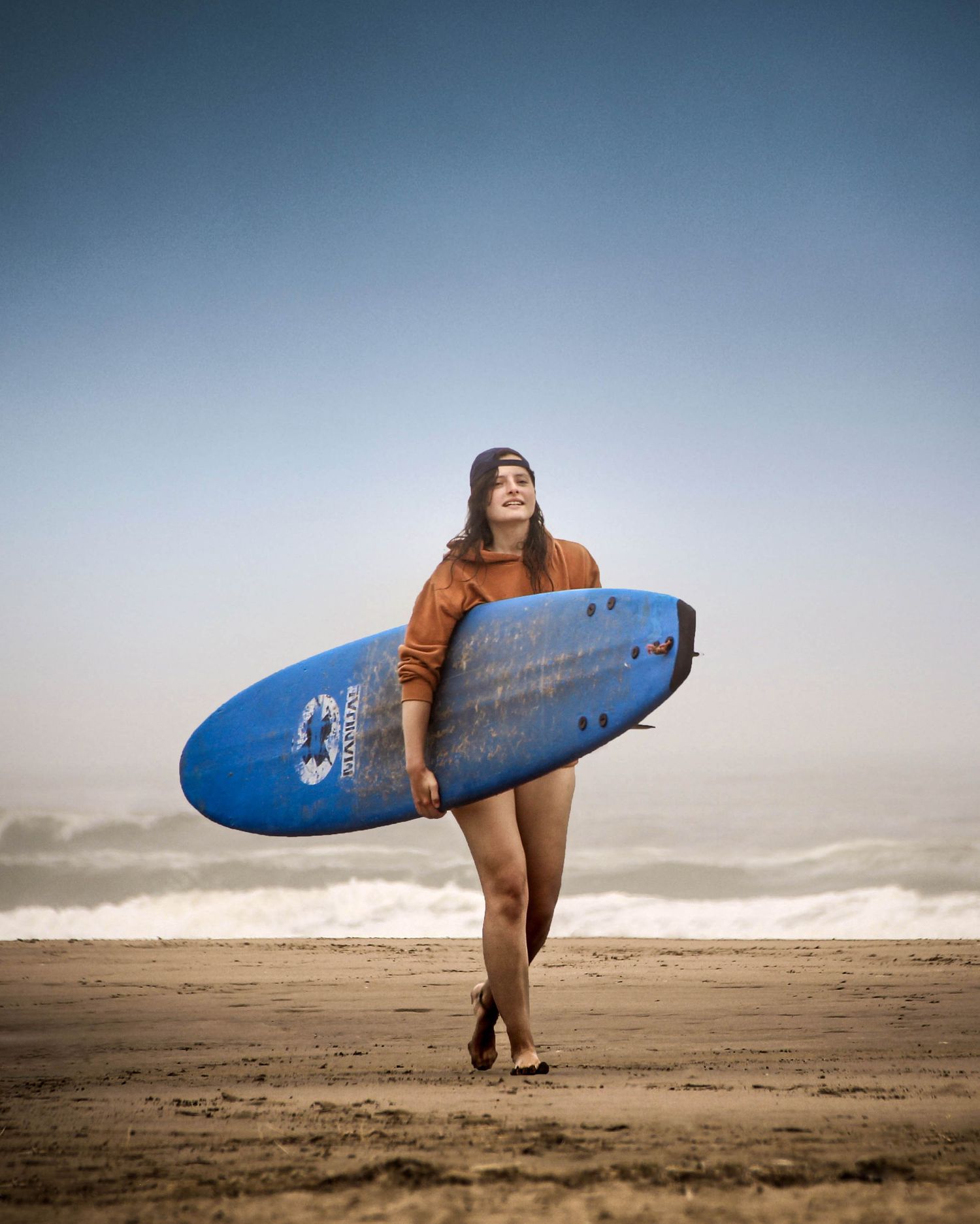 Woman in brown hoodie and beanie carries blue surfboard on foggy beach with waves in background.