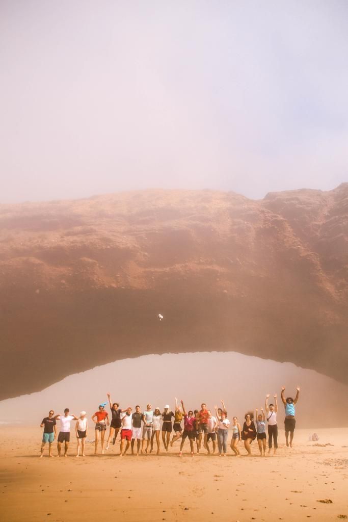 Group of people jumping and celebrating on a sandy beach with a dramatic rocky arch and misty mountains backdrop.