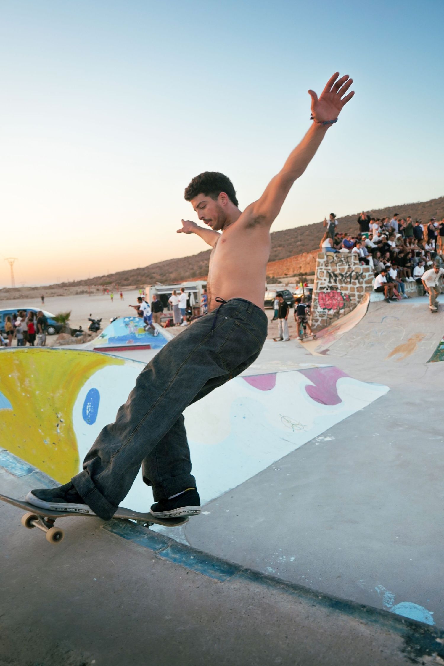 Shirtless skateboarder balancing on board with arms raised on a painted ramp at a crowded skate event.
