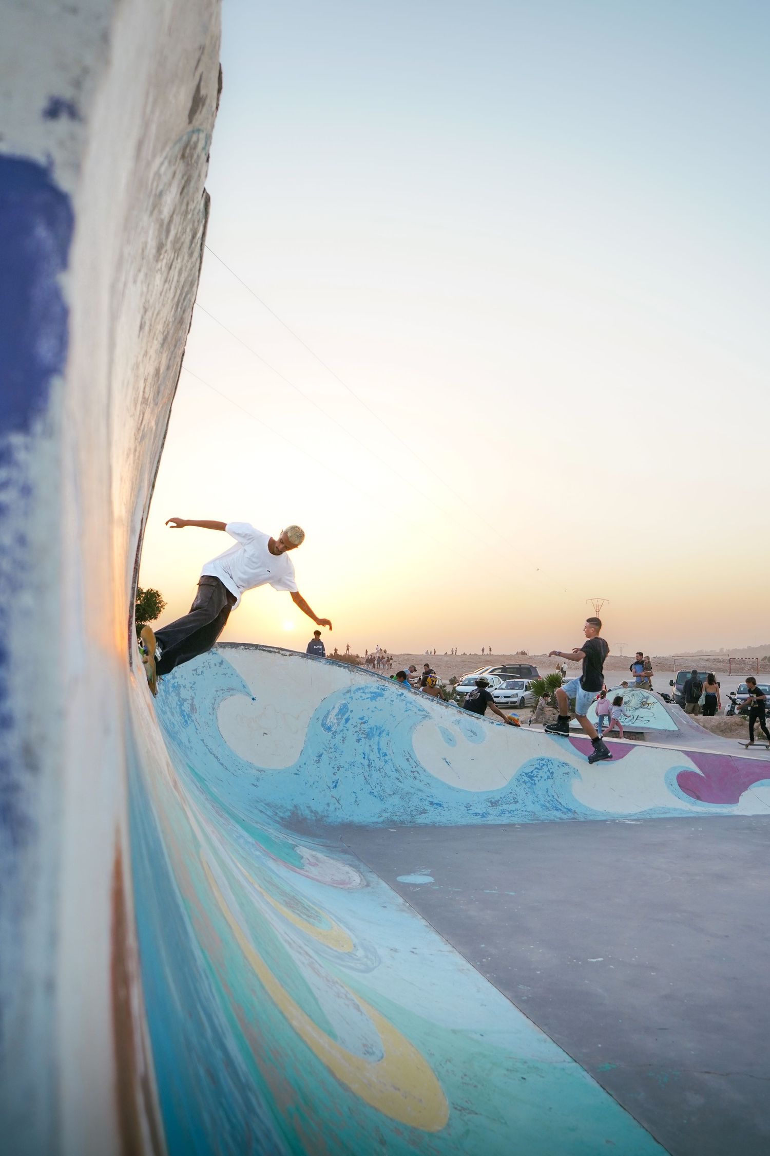Skateboarder performing wall ride trick at colorful outdoor skate park during golden sunset.