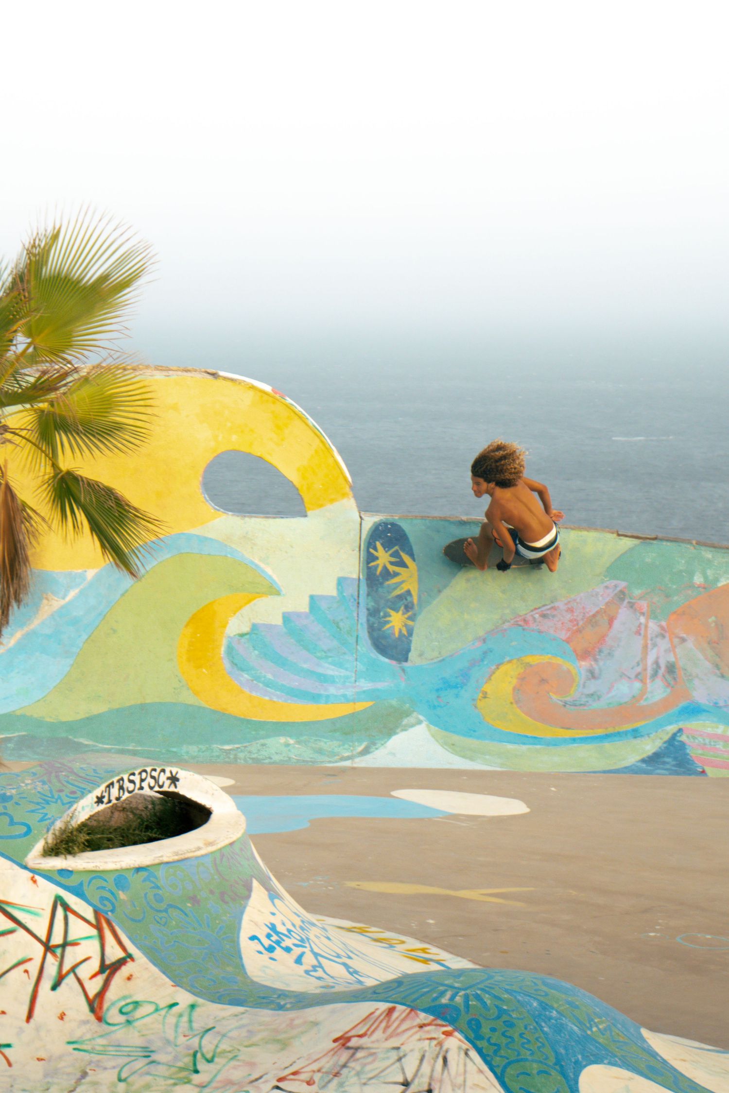 Child sitting on a colorful mural-painted skate ramp overlooking the ocean with a palm tree nearby.
